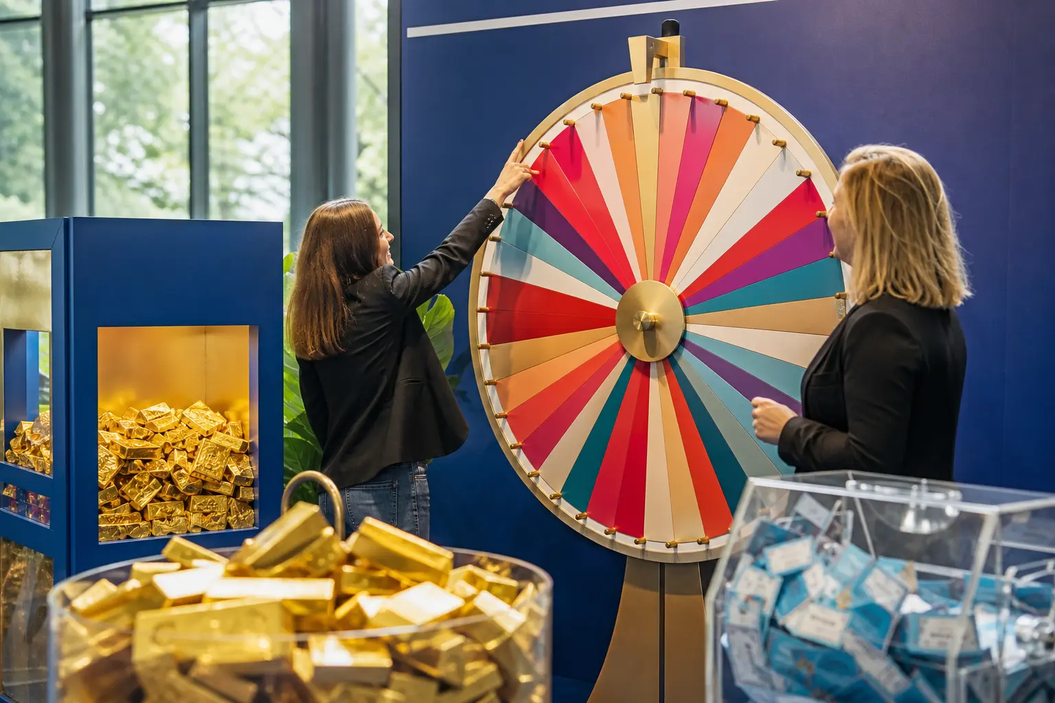 Deux femmes animent une roue de la fortune devant des lots en or, dans un espace commercial coloré.